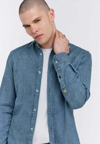 Camicia in denim blu chiaro con bottoni, maniche lunghe e colletto strutturato. Presenta un logo visibile sul polsino interno.