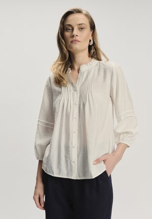 Blusa blanca de botones con cuello con volantes, pliegues verticales y mangas con puños. Tela ligera, diseño fluido, con dobladillo redondeado.