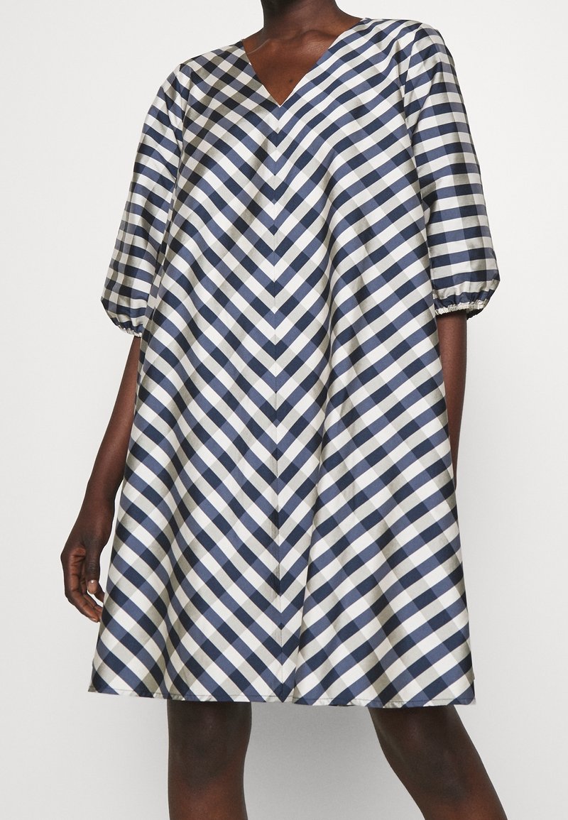Femme portant une robe longueur genou avec un motif à carreaux diagonaux bleu marine et blanc et des manches bouffantes aux coudes sur un fond uni.