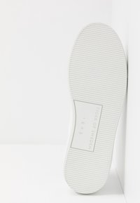 Bílá podrážka tenisky s texturovaným vzorem pro grip, s obdélníkovým logem, mírně zaobleným tvarem špičky a minimalistickým designem.