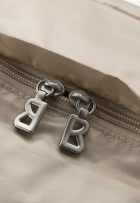 Béžová nylonová taška s hladkým povrchem, se dvěma stříbrnými zipy tvarovanými jako písmeno 'B' pro snadné otevírání.