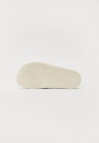 Semelle en caoutchouc blanche avec des motifs de grip texturés, présentant un bord lisse et un logo de marque moulé au talon. Design épuré et minimaliste.