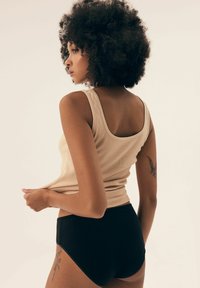 Beżowy top na ramiączkach z żebrowanej tkaniny i czarne figi z wysokim stanem. Gładka tekstura, dopasowany krój i tatuaż widoczny na udzie.