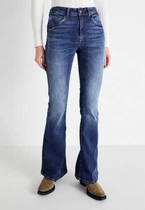 Kobieta w niebieskich spranych dżinsach bootcut i brązowych butach z kwadratowym noskiem, stojąca na tle jednolitego jasnego tła.