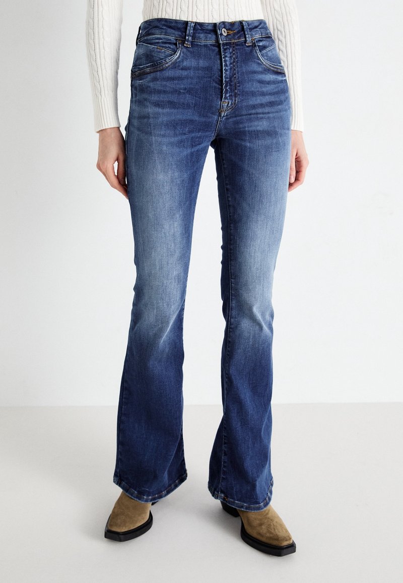Femme portant un jean bootcut bleu délavé et des bottes marron à bout carré, debout devant un fond clair uni.