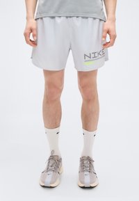 Ανοιχτό γκρι αθλητικό σορτς με μαύρο και νέον πράσινο λογότυπο Nike, με χαλαρή εφαρμογή και ελαστική μέση. Φορεμένο με λευκές κάλτσες μέχρι τον αστράγαλο και αθλητικά παπούτσια.