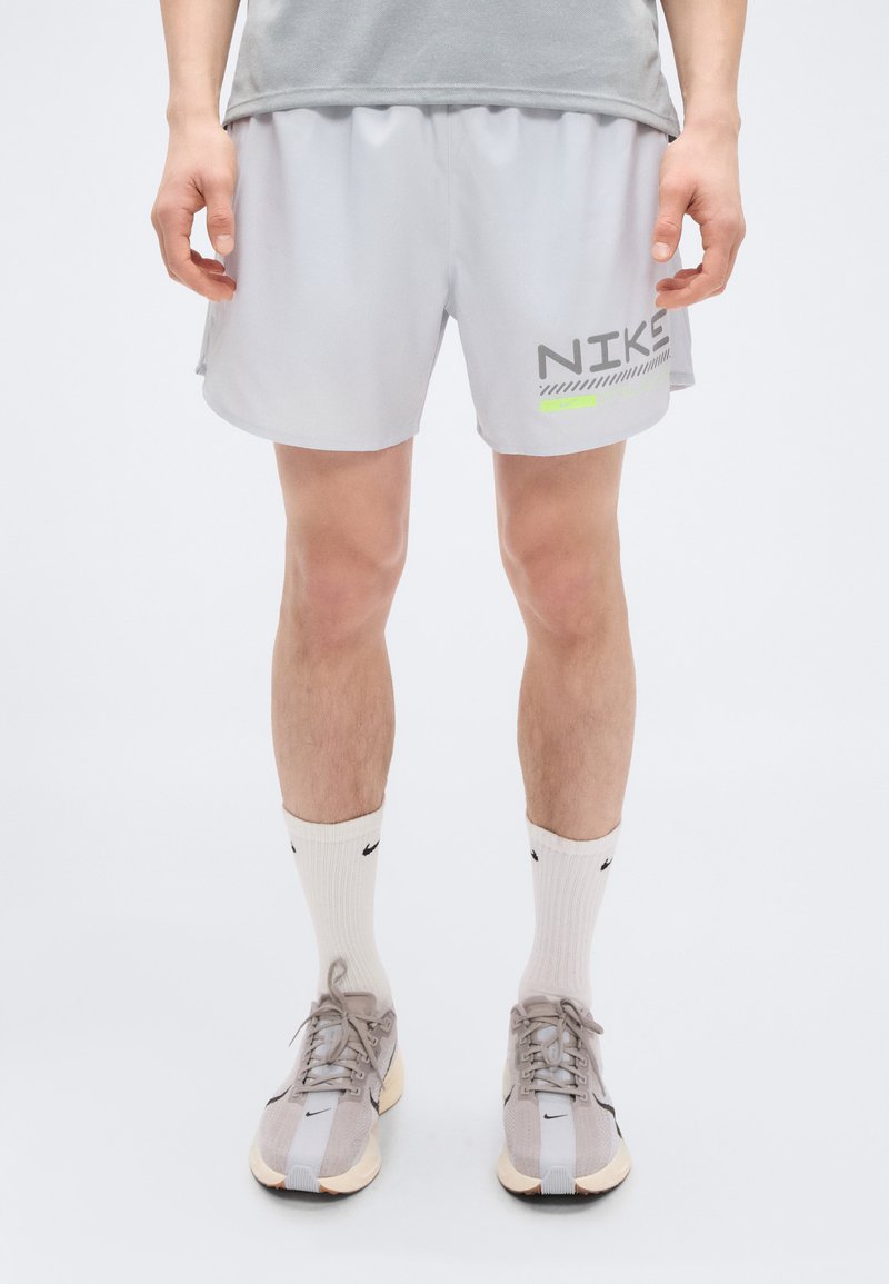 Ανοιχτό γκρι αθλητικό σορτς με μαύρο και νέον πράσινο λογότυπο Nike, με χαλαρή εφαρμογή και ελαστική μέση. Φορεμένο με λευκές κάλτσες μέχρι τον αστράγαλο και αθλητικά παπούτσια.