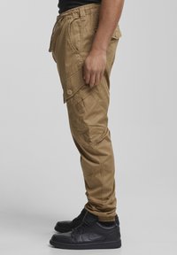 Kaki cargo hlače z elastičnim pasom, stranskimi žepi in sproščenim krojem. Nošene s črnimi usnjenimi supergami. Gladek tekstura, minimalni detajli.