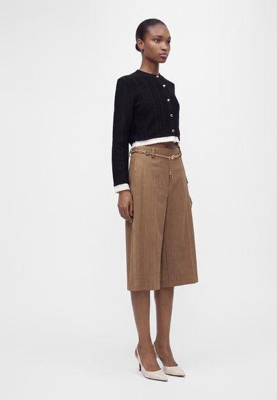 Femme debout portant un pantalon large court couleur camel avec ceinture chaîne dorée, veste courte noire avec poignets blancs et chaussures blanches à talons hauts.