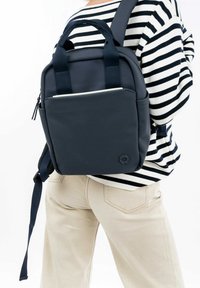 Námořnický batoh s dvojitými uchy, nastavitelnými popruhy, hladkým povrchem a přední kapsou, nošený na pruhovaném tričku a béžových kalhotách.