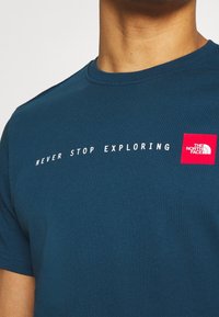 Modré tričko s bílým textem "NEVER STOP EXPLORING" a červeným čtvercovým logem značky The North Face na horní části hrudi.