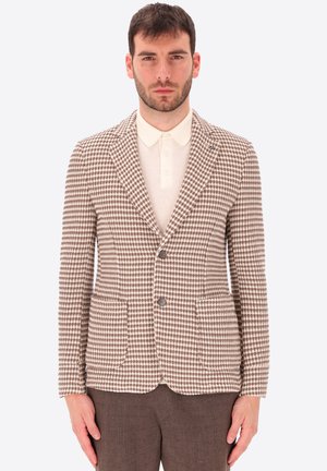 Uomo che indossa un blazer marrone e bianco a quadri sopra una polo crema e pantaloni marroni, in piedi su uno sfondo semplice.