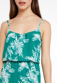 Grön ärmlös topp med vita bladmönster, med en draperad design och spagettiband. Mjuk tyg med en slät struktur.