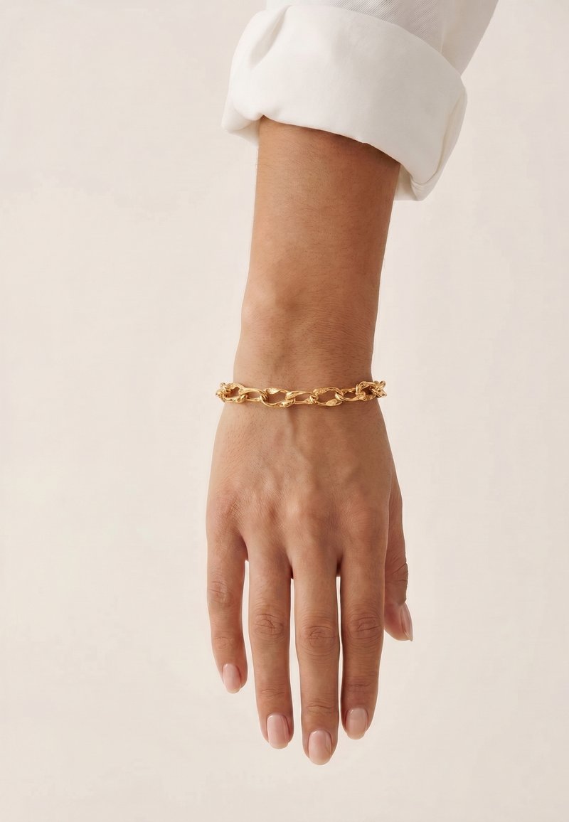 Poignet portant un bracelet chaîne torsadée en or, avec une manche de chemise blanche retroussée sur un fond clair uni.