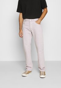 Hombre con jeans rosa claro, camiseta negra y zapatillas de lona beige, de pie con una mano en el bolsillo contra un fondo liso.