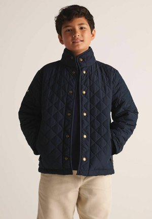 Junge trägt eine marineblaue gesteppte Jacke mit goldenen Knöpfen und beige Hose, steht mit den Händen in den Taschen vor einem schlichten Hintergrund.