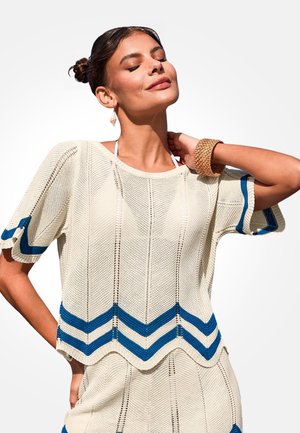 Frau mit geschlossenen Augen, die ein cremefarbenes gestricktes Oberteil mit blauen Chevron-Streifen und einen passenden Rock trägt, dazu Armband und Ohrring.