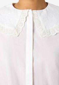Biała koszula z koronkowym, haftowanym kołnierzem. Tkanina jest jasnoróżowa, z frontowym zapięciem na guziki i delikatnym ażurowym wykończeniem.