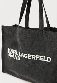 Fekete farmer szatyor dupla pánttal, melyen kihangsúlyozott fehér hímzett felirat található: "KARL LAGERFELD JEANS." Egyszerű téglalap alakú, masszív anyagból.