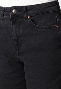 Lähikuva mustista farkuista, joissa on etutasku, vaskirivetti ja napin kiinnitys, yhdistettyinä valkoiseen ribbattuun yläosaan.