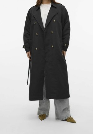 Woman wearing black double-breasted long coat, white textured top, light gray wide-leg jeans, and pointed olive green shoes.