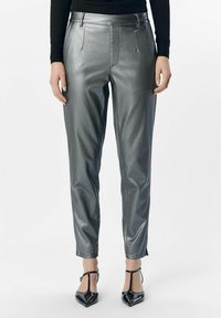 Metallische zilveren broek met een slim fit, een gladde textuur, voorplooien en ritsen bij de enkels. Gekleed met zwarte pumps met een spitse neus.