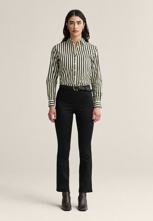 Nainen seisoo yllään mustat suoralahkeiset housut, musta vyö, ruskeat nilkkurit ja napitettu, pitkähihaisen paita, jossa on pystysuorat mustavalkoiset raidat.