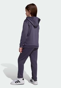 Girl standing sideways wearing a dark purple hoodie and pants with white Adidas sneakers on a light background.