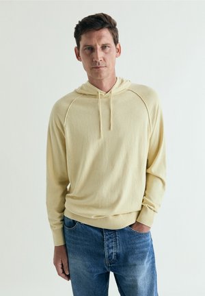 Hombre con suéter con capucha amarillo claro y jeans azules, de pie con una mano en el bolsillo contra un fondo liso.