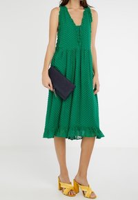 Grön klänning med polka prickar, V-ringning, volangdetaljer och utsvängd kjol. Matchad med en marinblå kuvertväska och gula plattformssandaler.