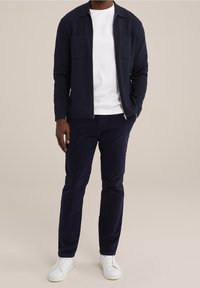 Man i marinblå zip-jacka, vit t-shirt, marinblå byxor och vita sneakers stående mot neutral bakgrund.