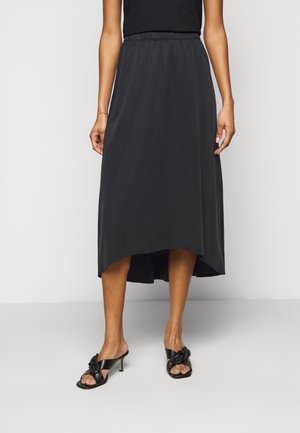 Femme portant une jupe noire courte devant et longue derrière, et des sandales noires à talons avec des brides nouées, debout devant un fond clair uni.