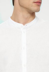 Άνδρας που φοράει λευκό λινό πουκάμισο με όρθιο γιακά και κάθετη σειρά κουμπιών, ορατός από το λαιμό έως το στήθος, κοιτάζει προς τα πλάγια.