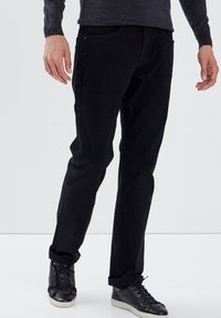Schwarze Cord-Hosen mit geradem Schnitt, ausgestattet mit vorderen Taschen und minimalen Hardware-Details. Kombiniert mit schwarzen Turnschuhen.