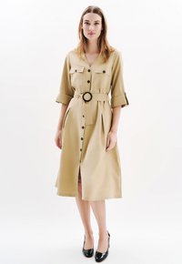 Vestito cammello con bottoni, maniche corte, cintura in vita, due tasche sul petto e uno spacco frontale. Presenta un tessuto liscio e strutturato.