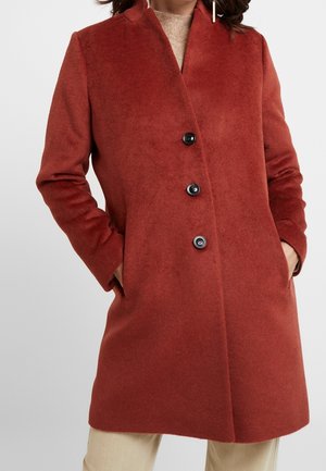 Femme portant un manteau en laine couleur rouille avec trois boutons noirs, mains dans les poches, pull beige et pantalon clair sur fond blanc.