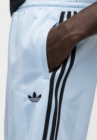 Αθλητικό παντελόνι ανοιχτό μπλε με μαύρες ρίγες στο πλάι και κεντημένο μαύρο λογότυπο. Το υλικό φαίνεται λείο με ελαφριά γυαλάδα.