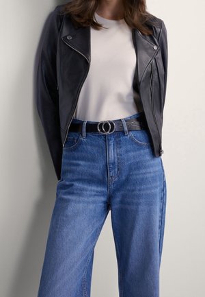 Kvinde iført sort læderjakke over hvid skjorte, der er stoppet ned i højtaljede blå jeans med sort bælte med dobbelt ring spænde.