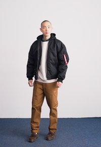 Black bomber jacket with zipper, hood, and red patch details, worn over a gray sweatshirt and brown pants, paired with dark shoes.