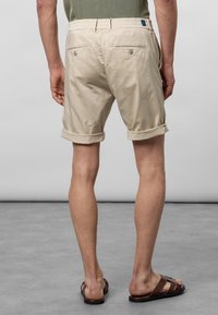 Shorts en coton beige à revers, dotés de deux poches arrière avec fermetures à bouton et d'une taille élastique. Portés avec des sandales marron.