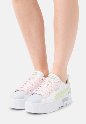 Jambes portant des baskets plateforme blanches Puma avec des lacets rose pâle et des bandes latérales vert clair sur un fond blanc.
