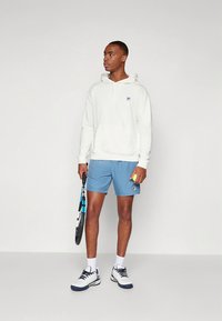 Bijela majica s kapuljačom, svijetloplave kratke hlače, drži tenisku lopticu i reket. Sportske tenisice s crnim i plavim detaljima. Jednostavan dizajn.