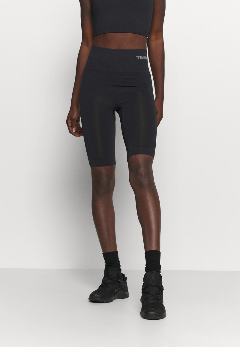 Personne portant un short cycliste taille haute noir, un crop top noir sans manches, des chaussettes noires et des chaussures de sport noires sur un fond uni.