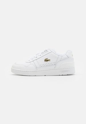 Witte sneaker van synthetisch leer met een platte zool, geperforeerde zijaccenten en een gouden Lacoste-logo aan de buitenkant.
