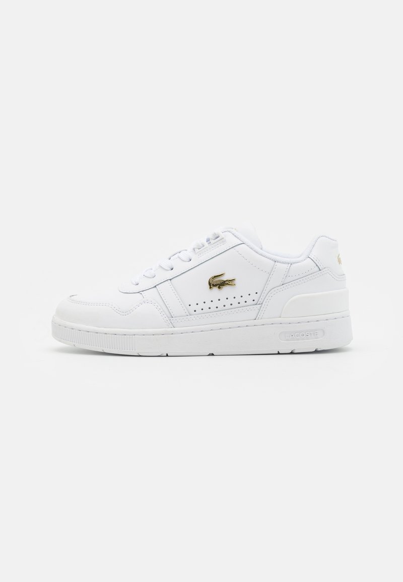 Baskets blanches en cuir synthétique avec une semelle plate, des détails latéraux perforés et un logo Lacoste doré sur le côté extérieur.