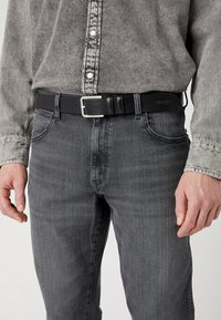 Zwarte leren riem met een rechthoekige zilveren gesp, gedragen over donkergrijze denim jeans en een grijs denim overhemd met knoopdetails.