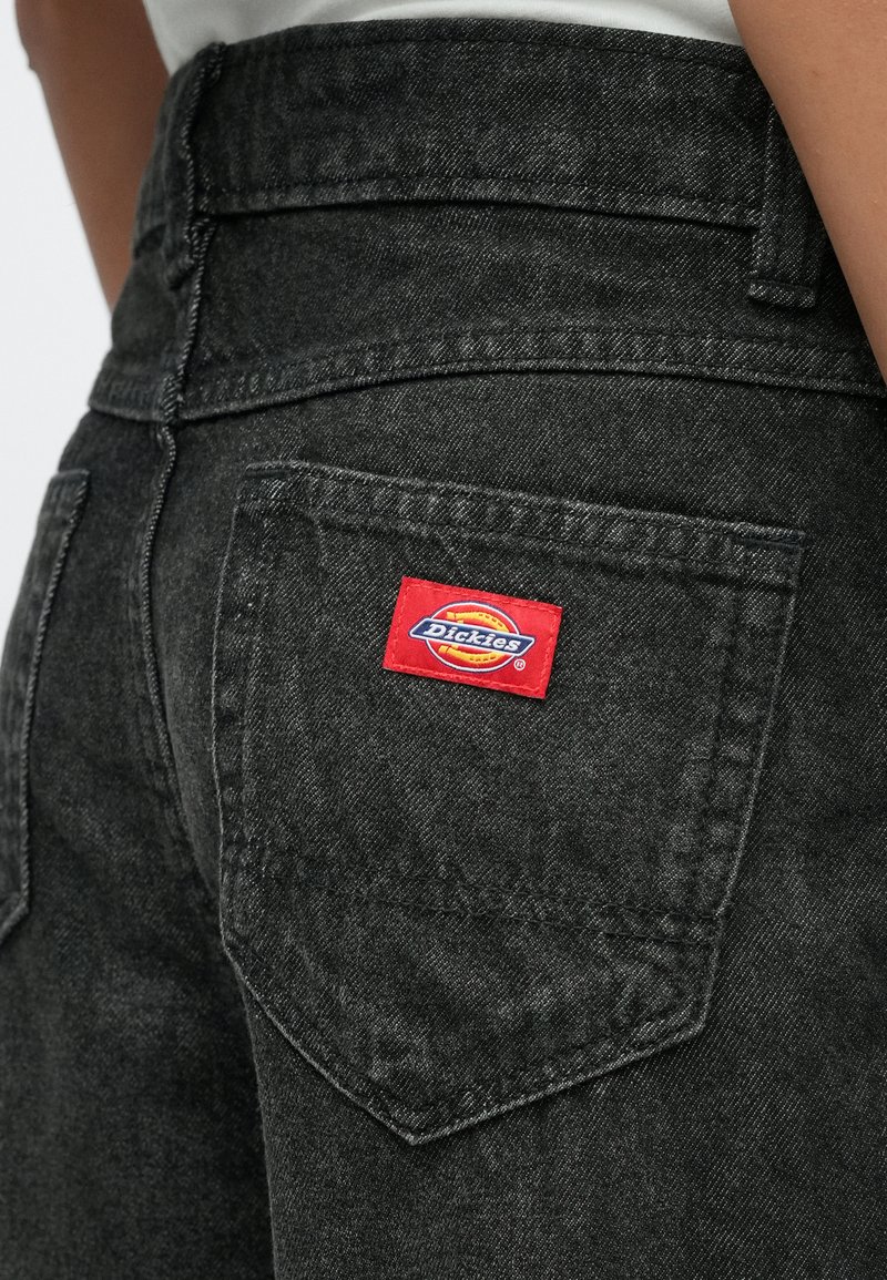 Schwarze Jeans-Tasche aus Denim mit aufgenähtem roten Dickies-Markenlabel, getragen von einer Person in weißem Hemd, Nahaufnahme der Hüftpartie.