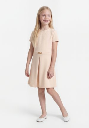 Młoda dziewczyna z długimi blond włosami uśmiechająca się, ubrana w jasno-różową sukienkę z krótkim rękawem i małym paskiem oraz białe baletki, stojąca na białym tle.