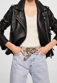 Giubbotto da motociclista in pelle nera con hardware argentato, indossato sopra una maglietta crema. Jeans in denim azzurro chiaro con cintura a serpente.