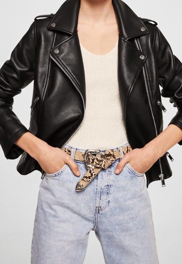 Giubbotto da motociclista in pelle nera con hardware argentato, indossato sopra una maglietta crema. Jeans in denim azzurro chiaro con cintura a serpente.